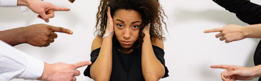 A woman standing up and covering here ears while people's fingers point at her.