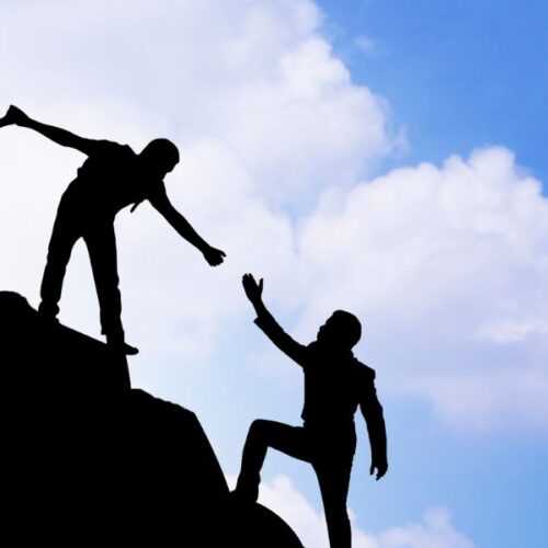 A group of people walking and helping each other up a cliff with a blue sky and clouds in the background.