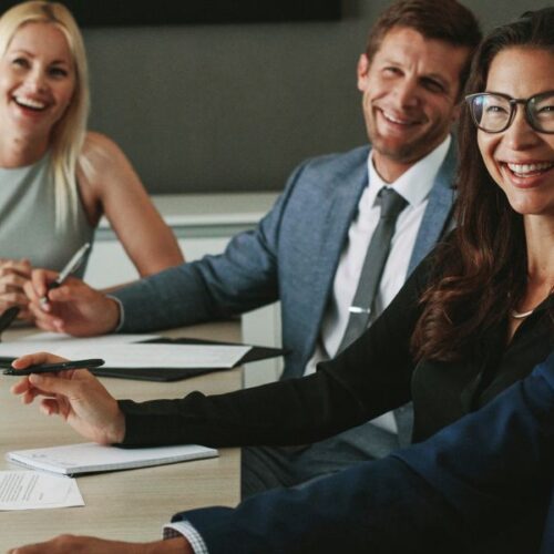 A group of employees sitting at a boardroom table smiling at an employee who is presenting.