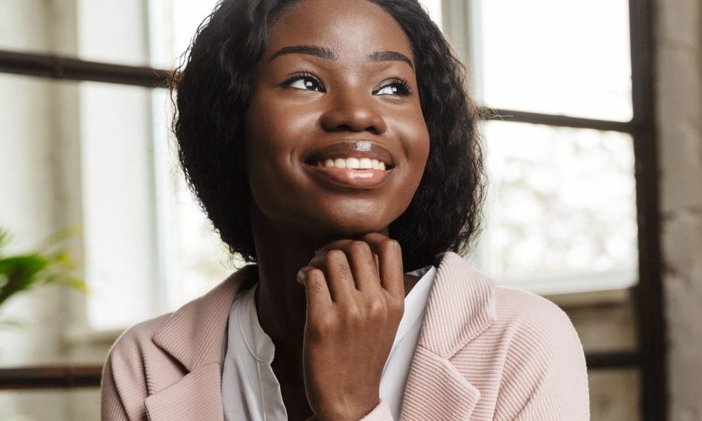 A woman sitting at a desk in an office resting her head on her chin and smiling.