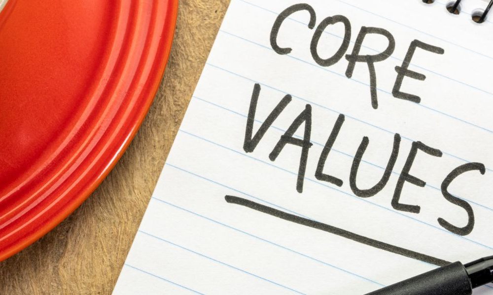 A red mug of coffee and a notepad that says Core Values with a pen on a light brown table.