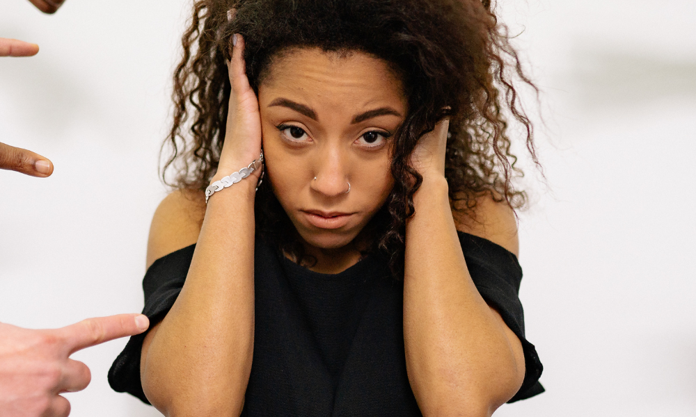 A woman standing up and covering here ears while people's fingers point at her.