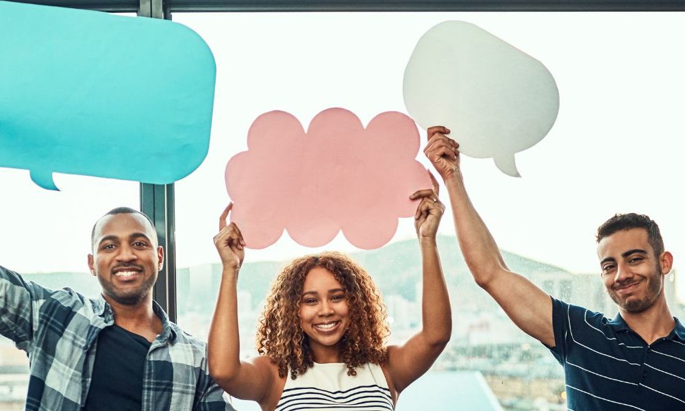 Five people standing in a line in front of an office window holding up paper cut-out speech bubbles.