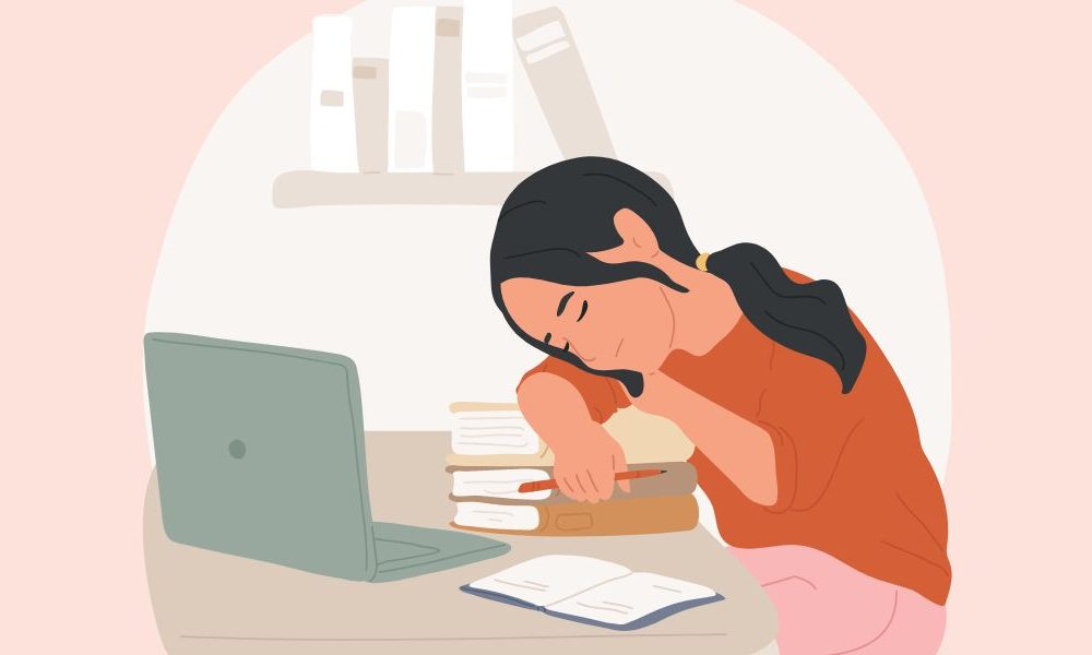 A woman at a desk resting her head on a stack of books in front of a laptop.