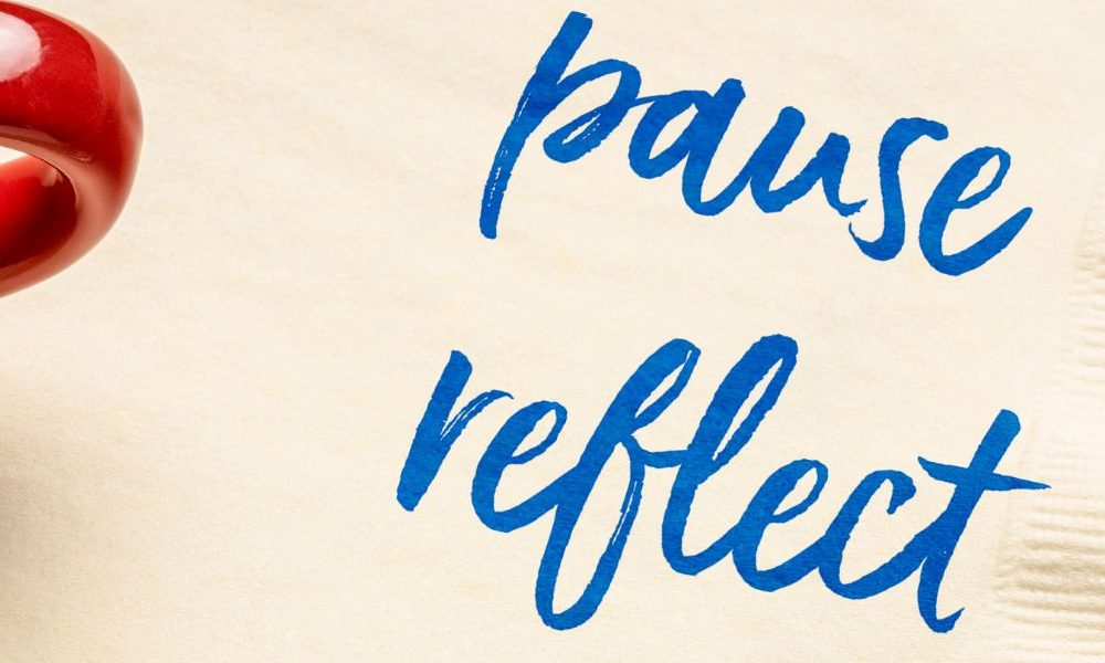 A napkin and cup of coffee on a table with the words Pause and Reflect written on the napkin.