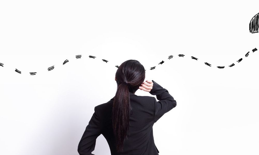 A woman in a suit looking at a drawing on a white wall of a squiggly dotted line connecting to a question mark.
