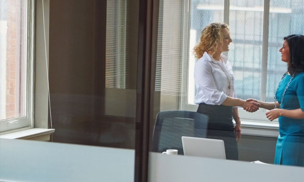 Two women shaking hands in a corporate office space.