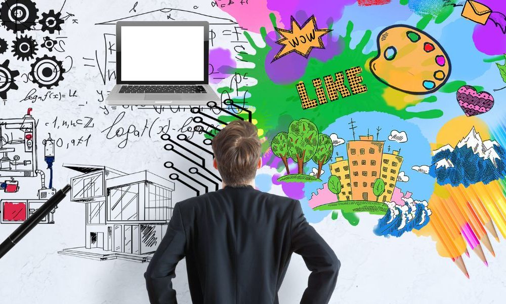 A man standing in front of a white wall with black and white industrial drawings to the left and colourful, creative drawings to the right.