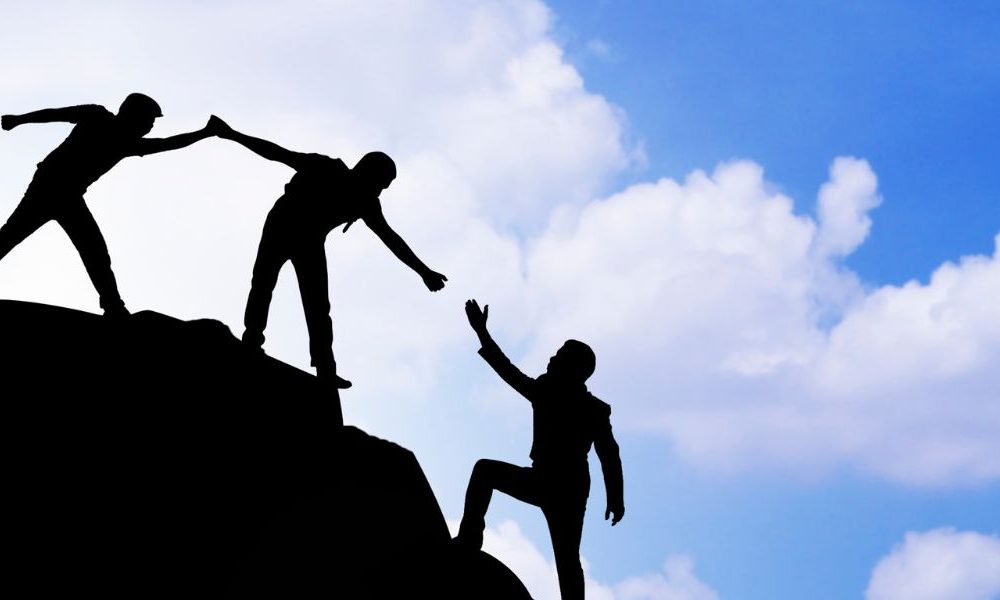 A group of people walking and helping each other up a cliff with a blue sky and clouds in the background.