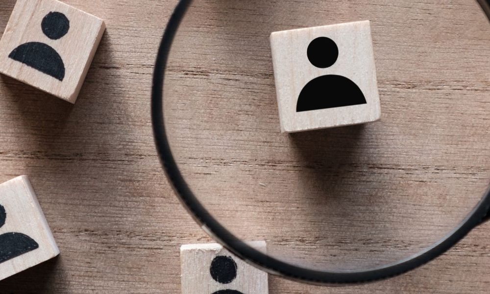 Wooden cubes with black outlines of people painted on them site on a table and someone is holding a magnifying glass over them.