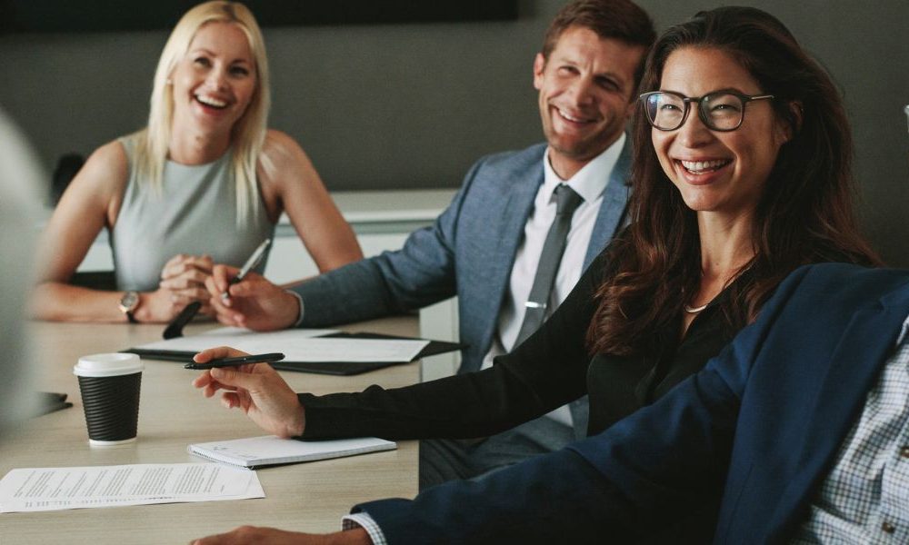 A group of employees sitting at a boardroom table smiling at an employee who is presenting.