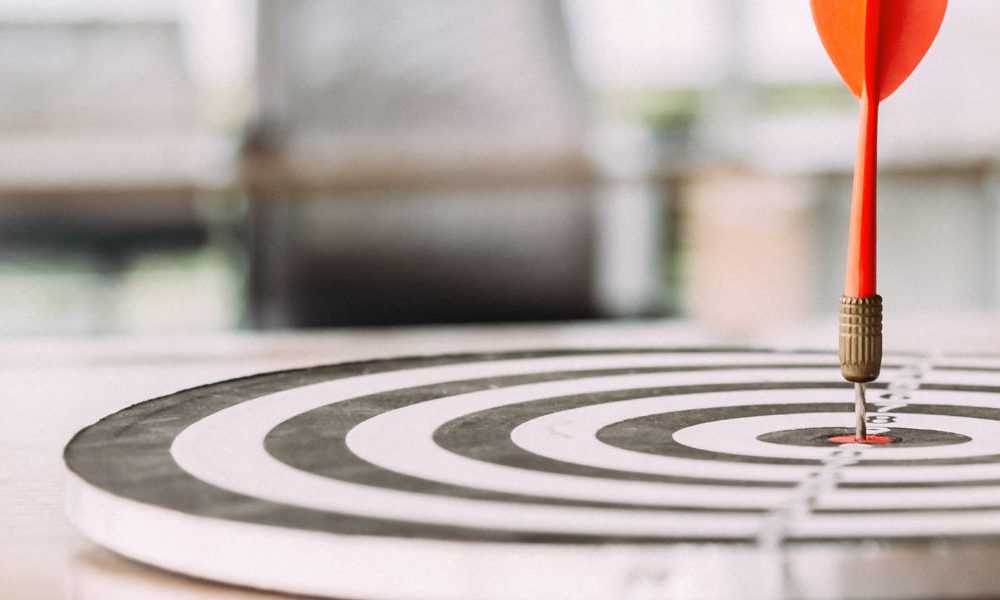 A dart in the centre of a dart board that's resting on a table in an office.