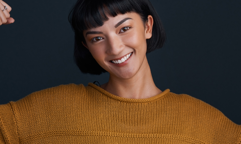 A woman wearing a mustard yellow sweater holding her arms in a muscle position in front of a dark grey background.