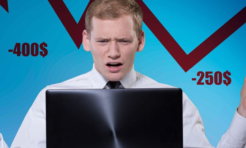 A man sitting in front of a laptop looking distressed with a blue background, downward arrow, and negative currency statements behind him.