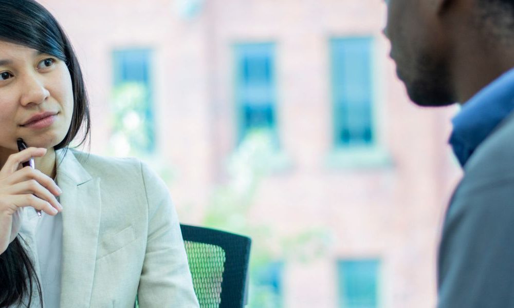 A woman sitting across a table from a man in an office while actively listening to his conversation.